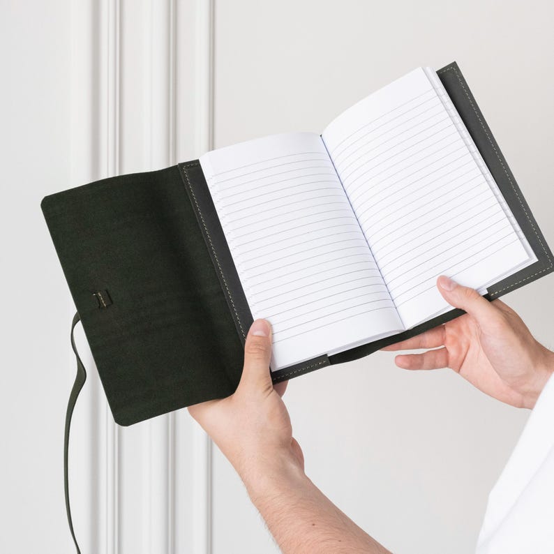 May include: A dark green leather notebook with a strap closure. The notebook is open to reveal lined pages.