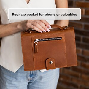 May include: A brown leather handbag with a front zippered pocket and a handle. The bag has a rectangular shape and a button closure. The text "Rear zip pocket for phone or valuables" is visible.