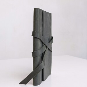 May include: A dark green leather journal with a leather strap closure. The strap is wrapped around the journal and tied in a knot.