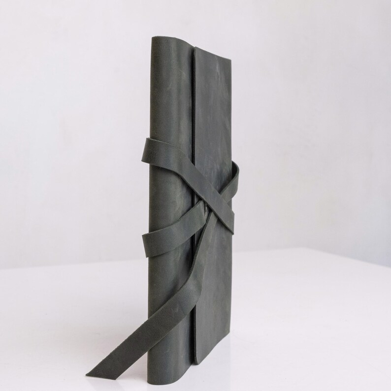 May include: A dark green leather journal with a leather strap closure. The strap is wrapped around the journal and tied in a knot.