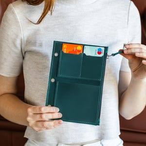 May include: A teal leather wallet with multiple card slots and a zippered compartment. The wallet is open, revealing credit cards. The wallet has a small loop attached to the zipper. The wallet is held by a person.