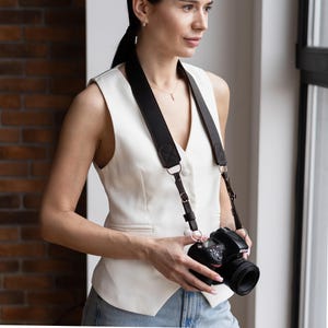 May include: Black leather camera strap with silver metal hardware. The strap is attached to a black camera with a lens. The camera strap is worn over the shoulder.