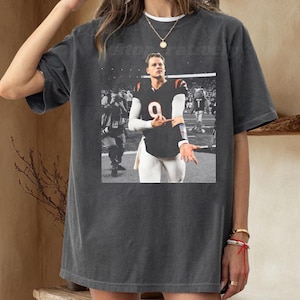 May include: Dark grey t-shirt with a black and white image of a football player in a black jersey with the number 9. The player is gesturing with his hands. The shirt has a relaxed fit.