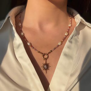 May include: A gold-toned necklace with a sunburst pendant. The necklace features a mix of small, colorful beads, freshwater pearls, and a central pendant with a starburst design and a central stone. The necklace is worn over a white collared shirt.