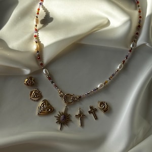 May include: A beaded necklace with pearls and colorful beads, along with several gold-colored pendants, including crosses, a heart, and a sunburst design. The jewelry is displayed on a white, silky fabric.