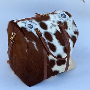 Puede incluir: Una bolsa de lona rectangular grande con un estampado de piel de vaca en marrón y blanco. La bolsa tiene asas de cuero marrón, un cierre de cremallera y una correa para el hombro desmontable. Los herrajes dorados acentúan la bolsa.
