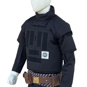 Pu&ograve; includere: Gilet tattico nero con maniche corte, collo alto e toppe rettangolari. Una cintura in pelle marrone con fibbia dorata e due tasche &egrave; indossata in vita. Il logo Galactic Props &egrave; visibile.