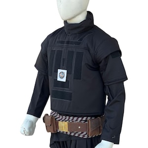 Pu&ograve; includere: Gilet tattico nero con maniche corte, collo alto e pi&ugrave; toppe rettangolari. Una cintura in pelle marrone con fibbia dorata e tasche &egrave; indossata in vita. L'outfit &egrave; esposto su un manichino.
