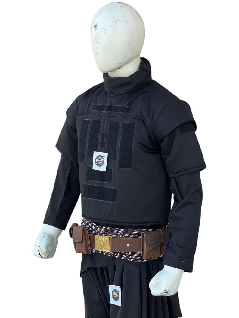 Pu&ograve; includere: Gilet tattico nero con maniche corte, collo alto e pi&ugrave; toppe rettangolari. Una cintura in pelle marrone con fibbia dorata e tasche &egrave; indossata in vita. L'outfit &egrave; esposto su un manichino.