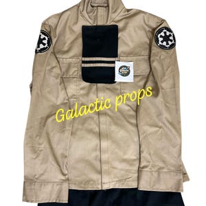 May include: Tan jacket with black accents, two front pockets, and a black rectangle on the chest. The jacket features two circular patches with a black and white design on the sleeves. The text "Galactic props" is written in yellow.