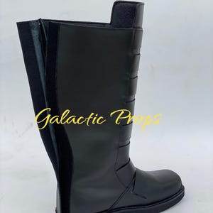 May include: A black knee-high boot with a flat sole. The boot features a side zipper, decorative straps, and the text "Galactic Props" in gold script. The boot is made of a smooth, matte material.