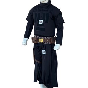 Pu&ograve; includere: Un costume nero esposto su un manichino. L'outfit include un top a maniche corte con collo alto, un gilet con pannelli rettangolari e pantaloni a gamba larga. Una cintura marrone con fibbia dorata e una fondina completano l'ensemble.