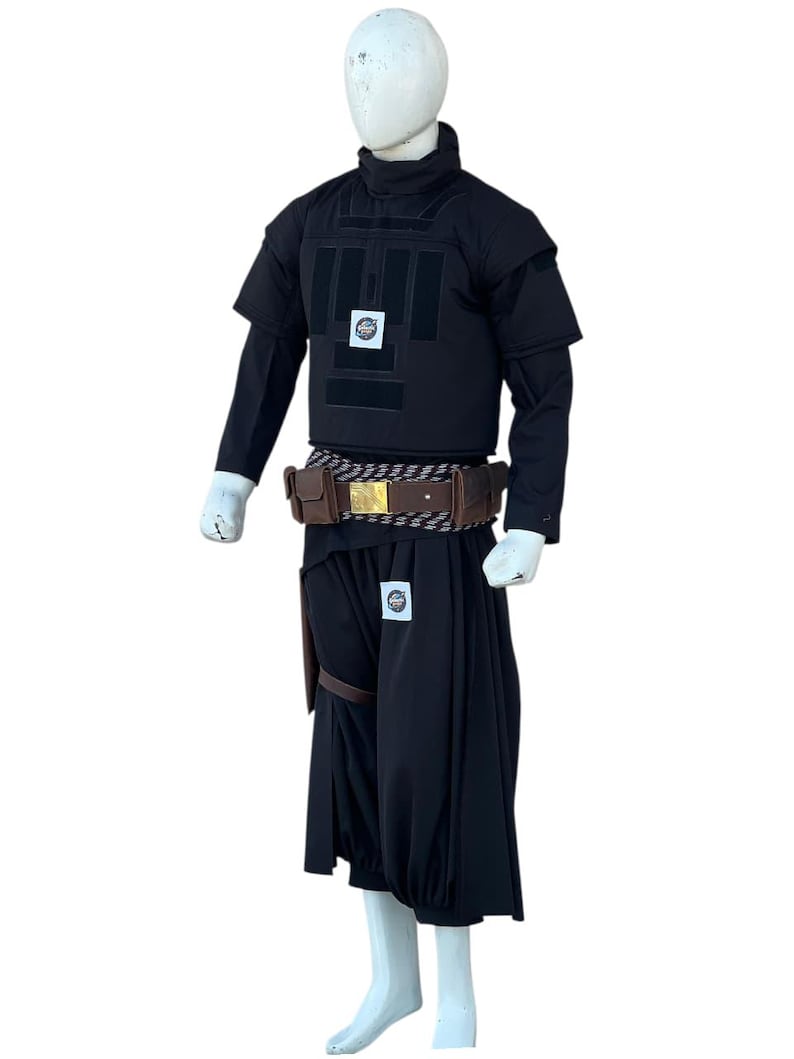 Pu&ograve; includere: Un costume nero esposto su un manichino. L'outfit include un top a maniche corte con collo alto, un gilet con pannelli rettangolari e pantaloni a gamba larga. Una cintura marrone con fibbia dorata e una fondina completano l'ensemble.