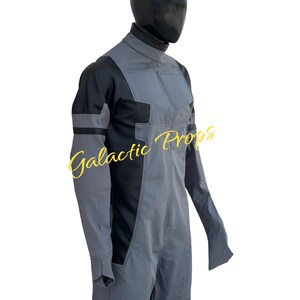 May include: A gray and black jumpsuit with a high collar and long sleeves. The jumpsuit has a textured pattern and is displayed on a mannequin. The text "Galactic Props" is visible in yellow script.