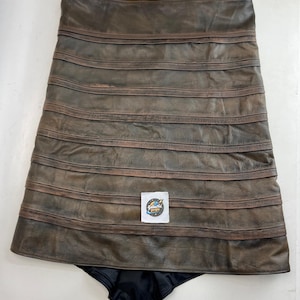 Könnte beinhalten: Ein brauner Lederrock mit horizontalen Bändern und dunklem Futter. Der Rock hat ein abgenutztes Aussehen und ein Etikett mit der Aufschrift "Galactic Props".