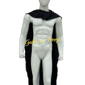 Può includere: Un mantello nero drappeggiato su un manichino bianco. Il mantello è fatto di un tessuto scuro e scende lungo la schiena del manichino. Il manichino è su uno sfondo bianco. Il testo sul manichino recita "Galactic Props".