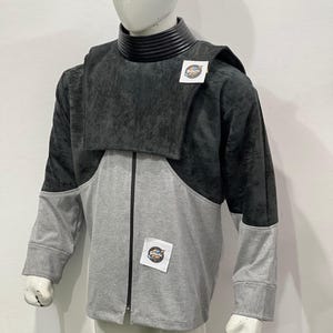 May include: A two-tone jacket on a mannequin. The upper portion is dark gray, with a textured appearance, and the lower section is light gray. A black collar and a zipper are visible. The jacket has a patch that says "Galactic Props".