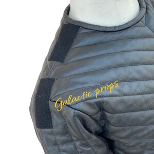 Puede incluir: Chaqueta de cuero acolchada negra con correas de velcro negras en los hombros. El texto "Galactic props" est&aacute; impreso en amarillo en la chaqueta.