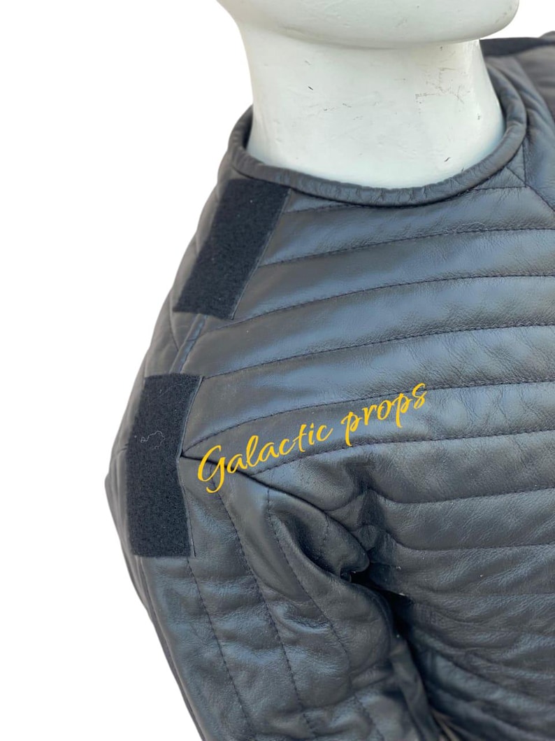 Puede incluir: Chaqueta de cuero acolchada negra con correas de velcro negras en los hombros. El texto "Galactic props" est&aacute; impreso en amarillo en la chaqueta.