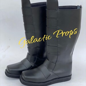 May include: A pair of black knee-high boots with a square toe and multiple horizontal straps. The boots have a smooth, shiny finish and are likely made of leather or a leather-like material. The text "Galactic Props" is visible in yellow.