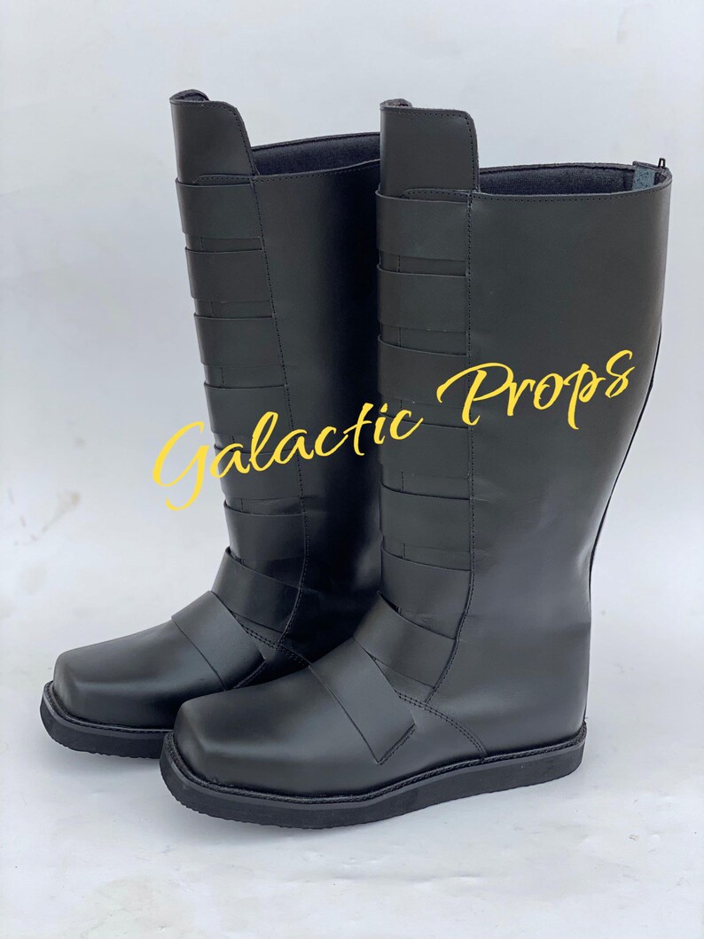 May include: A pair of black knee-high boots with a square toe and multiple horizontal straps. The boots have a smooth, shiny finish and are likely made of leather or a leather-like material. The text "Galactic Props" is visible in yellow.