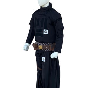 Pu&ograve; includere: Completo tattico nero, che include un gilet con pi&ugrave; pannelli, una camicia a maniche lunghe e pantaloni fluidi simili a una gonna. Sono visibili anche una cintura marrone con tasche e un cinturino in pelle. L'outfit &egrave; esposto su un manichino.