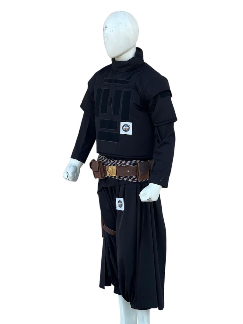 Pu&ograve; includere: Completo tattico nero, che include un gilet con pi&ugrave; pannelli, una camicia a maniche lunghe e pantaloni fluidi simili a una gonna. Sono visibili anche una cintura marrone con tasche e un cinturino in pelle. L'outfit &egrave; esposto su un manichino.