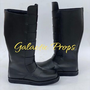 May include: A pair of black knee-high boots. The boots have a smooth, matte finish and feature several horizontal straps across the upper shaft. The text "Galactic Props" is printed in yellow cursive on the boots.
