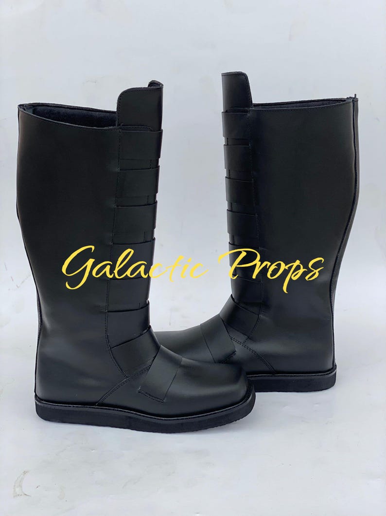 May include: A pair of black knee-high boots. The boots have a smooth, matte finish and feature several horizontal straps across the upper shaft. The text "Galactic Props" is printed in yellow cursive on the boots.