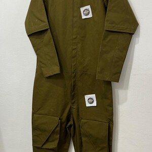 May include: Olive green jumpsuit with a zip-up front, high collar, and multiple pockets. Features patches on the chest and a relaxed fit. The jumpsuit is hanging on a metal hanger.