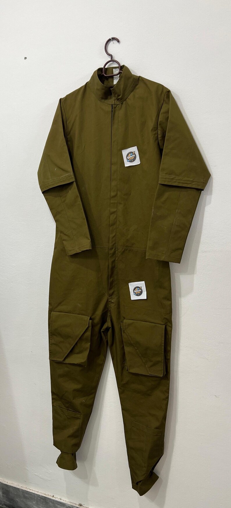 May include: Olive green jumpsuit with a zip-up front, high collar, and multiple pockets. Features patches on the chest and a relaxed fit. The jumpsuit is hanging on a metal hanger.