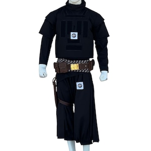Pu&ograve; includere: Abito tattico nero su manichino. L'abito comprende un gilet, una camicia a maniche lunghe e pantaloni larghi. Sono inclusi anche una cintura marrone con fibbia dorata e una fondina in pelle. Toppe con un logo circolare sono visibili sul gilet e sui pantaloni.