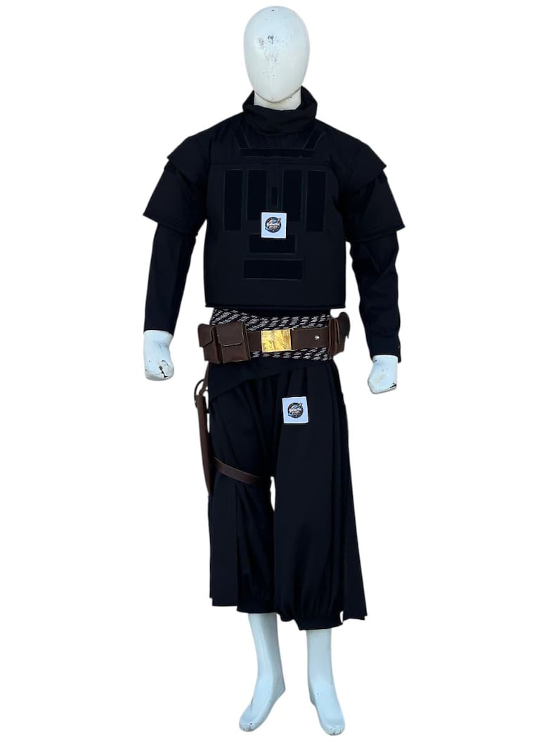 Pu&ograve; includere: Abito tattico nero su manichino. L'abito comprende un gilet, una camicia a maniche lunghe e pantaloni larghi. Sono inclusi anche una cintura marrone con fibbia dorata e una fondina in pelle. Toppe con un logo circolare sono visibili sul gilet e sui pantaloni.