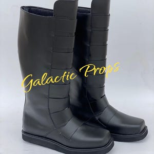 May include: A pair of black knee-high boots with a square toe and multiple horizontal straps. The boots have a smooth, leather-like appearance and a flat sole. The text "Galactic Props" is written in yellow script across the image.