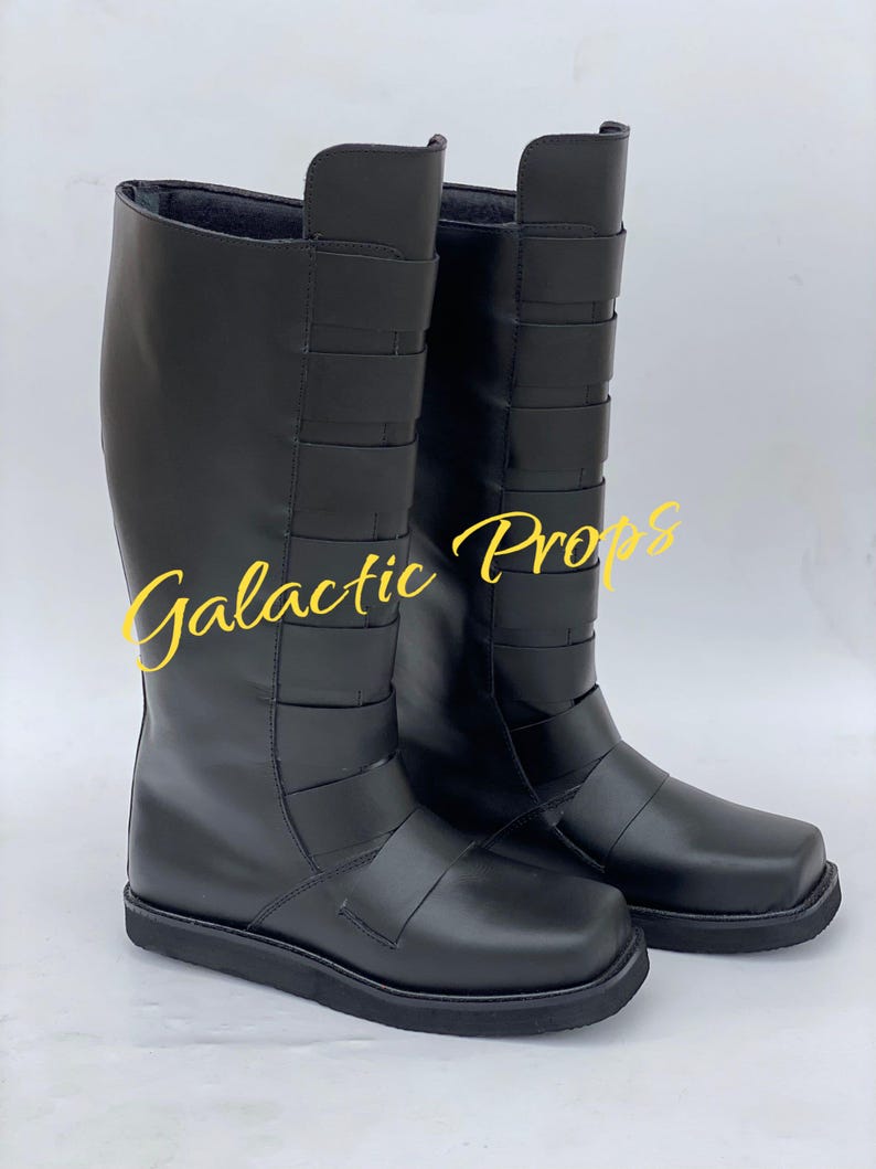 May include: A pair of black knee-high boots with a square toe and multiple horizontal straps. The boots have a smooth, leather-like appearance and a flat sole. The text "Galactic Props" is written in yellow script across the image.