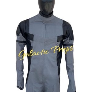 May include: A gray and black jumpsuit with a high collar and long sleeves. The jumpsuit has a textured pattern on the front and sleeves, with black panels on the shoulders and sides. The text "Galactic Props" is written in a cursive font.