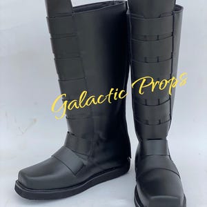May include: Pair of black knee-high boots with multiple straps and a square toe. The boots have a smooth, matte finish and are displayed on a white surface. The text "Galactic Props" is visible in the image.