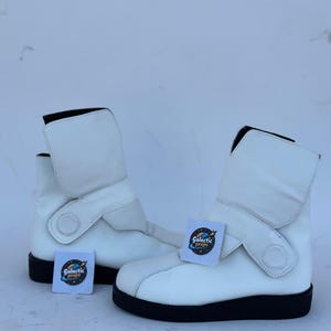Könnte beinhalten: Ein Paar weiße Stiefel mit schwarzen Sohlen. Die Stiefel haben eine hochklappbare Klappe, die mit einem Riemen und einem kreisförmigen Knopf befestigt ist. Jeder Stiefel hat ein Etikett mit der Aufschrift "Galactic Props".