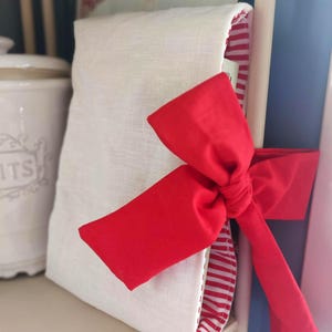 Può includere: Una borsa di lino bianco legata con un fiocco rosso. L'interno della borsa presenta un motivo a righe rosse e bianche. La borsa è esposta su una mensola bianca, con un'insegna vintage sullo sfondo.