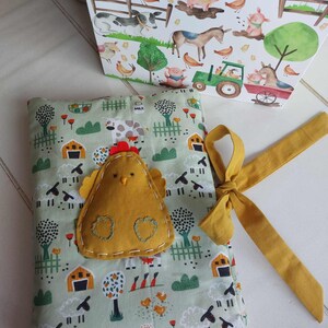 May include: A farm-themed fabric book with a yellow felt chicken applique and a mustard yellow ribbon. The book features a light green background with sheep, barns, and other farm animals. A matching box with farm animal illustrations is also visible.