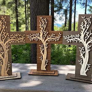 May include: Three wooden crosses with tree-of-life designs. Each cross is a different shade of brown and has a light-colored tree design cut out of the center. The crosses are on wooden bases and have Bible verses etched into them.