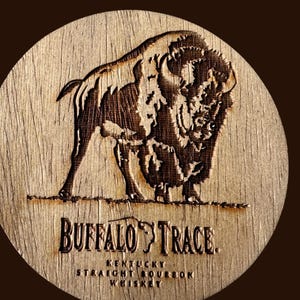 May include: A round wooden coaster with a detailed engraving of a bison. The coaster features the text "BUFFALO TRACE" and "KENTUCKY STRAIGHT BOURBON WHISKEY" in a serif font. The coaster is light brown with darker brown details.