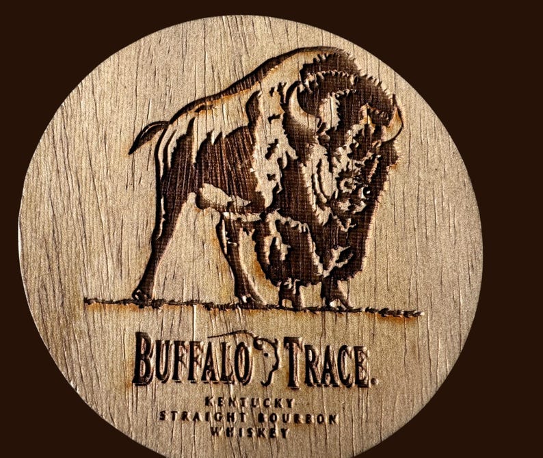 May include: A round wooden coaster with a detailed engraving of a bison. The coaster features the text "BUFFALO TRACE" and "KENTUCKY STRAIGHT BOURBON WHISKEY" in a serif font. The coaster is light brown with darker brown details.
