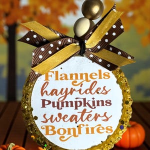May include: A seasonal ornament with a white circular centre displaying the words "Flannels, hayrides, Pumpkins, sweaters, Bonfires" in orange and brown. The ornament has a gold glitter border, a brown and white polka dot bow, and gold beads. Two small orange pumpkins are in the foreground.