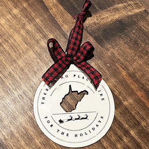 May include: Round white Christmas ornament with a cutout of the state of West Virginia. The ornament has the text "THERE'S NO PLACE LIKE FOR THE HOLIDAYS" and a red and black plaid ribbon.