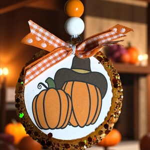 May include: A circular ornament with a pumpkin and cowboy hat design, edged with glitter. It has an orange and white gingham ribbon bow and is suspended by a white cord with orange and white beads. The background features blurred pumpkins.