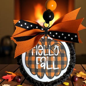May include: A round, pumpkin-shaped ornament with the words "HELLO FALL" in white. The ornament has a black and orange plaid design, a black and white polka dot bow, and a black beaded trim. An orange and black beaded string is attached.