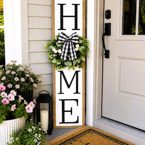 May include: A tall rectangular wooden porch sign with a white background and black lettering spelling HOME. The letter O is replaced by a green artificial wreath adorned with a black and white buffalo check ribbon bow, centred on the sign.