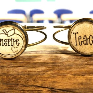 Op de afbeelding: Twee gouden armbandjes met ronde houten bedels. De bedel aan de linkerkant heeft een rode appel met het woord "inspireren" en een klein hartje. De bedel aan de rechterkant heeft het woord "leren" erop.