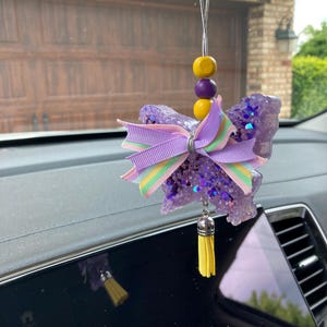 May include: Purple and yellow car air freshener with a glitter finish and a bow. The air freshener is shaped like a butterfly and has a tassel.